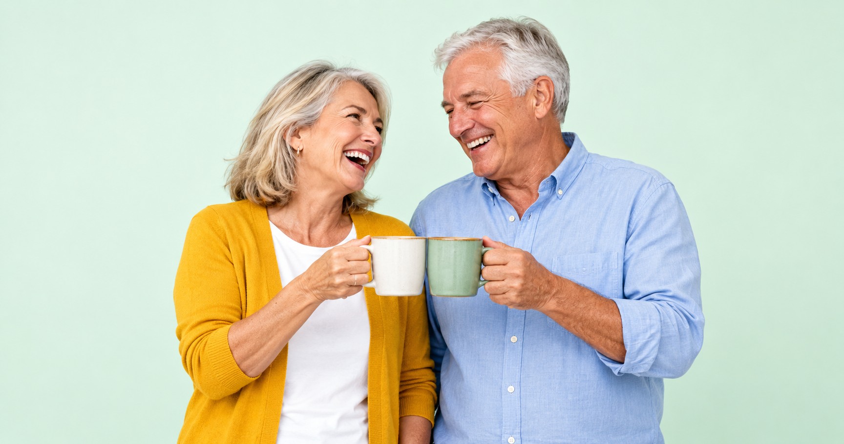 A laughing couple in their mid-sixties clinking coffee mugs against a soft mint background.