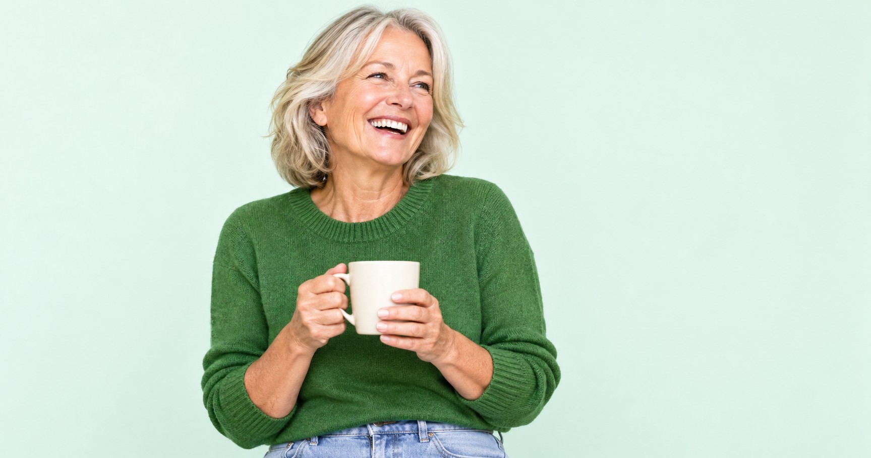 A smiling woman in her early sixties in a green sweater, holding a mug against a soft mint background.