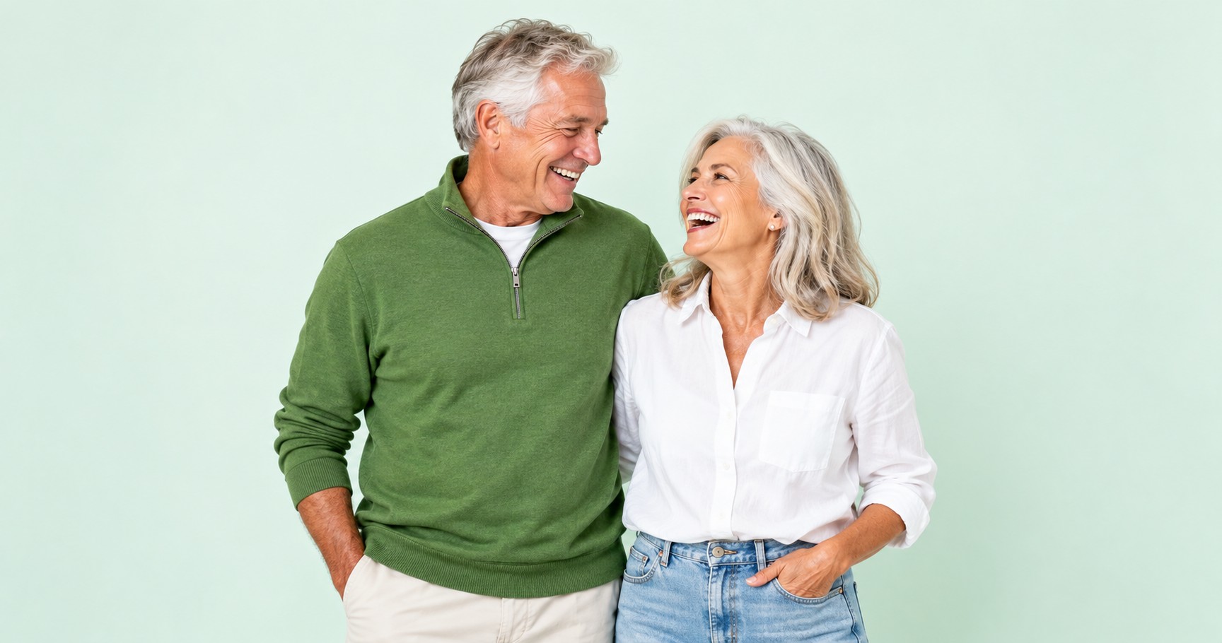A laughing couple in their sixties standing shoulder to shoulder against a soft mint background.